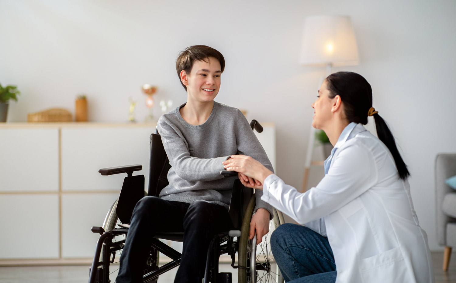 Disabled people healthcare support. Doctor holding hands of handicapped teen boy in wheelchair during medical visit